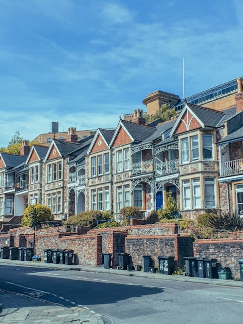 A row of Victorian-style terraced houses with bay windows, decorative white iron railings, and small front gardens enclosed by low brick walls, situated beside a paved street lined with black wheelie bins, with a clear blue sky overhead. This residential setting showcases typical urban housing, which may require professional house removals or home relocation services. The image emphasizes the exterior environment where a man with van service, such as Man with Van Wealdstone, could assist with furniture transport and packing during a local move. The houses are positioned along a slight incline, with some front gardens featuring bushes and hedges, and the street appears quiet and well-maintained, suitable for loading and unloading moving vehicles.