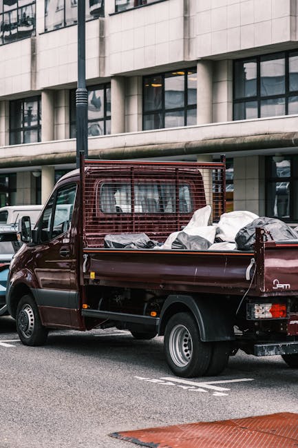 A small burgundy flatbed van parked on a city street near a modern office building with large glass windows and concrete panels. The van's cargo area is filled with various household items wrapped in plastic and fabric, including cardboard boxes, furniture cushions, and covered appliances. The items are arranged for transport, with some secured by straps, indicating an ongoing loading or unloading process. The vehicle is positioned close to the curb, with the rear and side doors open to facilitate the movement of belongings, suggesting a home relocation or furniture transport. The scene is illuminated with natural daylight, emphasizing the urban environment and the practical aspect of house removals, as managed by companies like Man with Van Wealdstone.