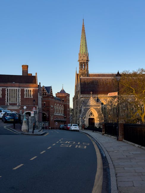 The image displays a narrow street in Wealdstone with several parked cars along the pavement. In the background, there is a prominent historic church with a tall, pointed spire, constructed from light-colored stone and featuring gothic architectural elements such as arched windows and decorative stonework. To the left of the church, there are red-brick buildings, some with bay windows and chimneys, typical of the area's residential and commercial architecture. The street is quiet, illuminated by soft natural light, suggesting early morning or late afternoon. A street lamp and traffic signage are visible, with a bus stop marking the edge of the road. Inside this setting, [COMPANY_NAME], a professional removals service, could be involved in a home relocation, furniture transport, or packing and moving process, possibly preparing for a house move in Wealdstone. The scene provides a clear view of the street environment relevant to house removals and moving logistics.
