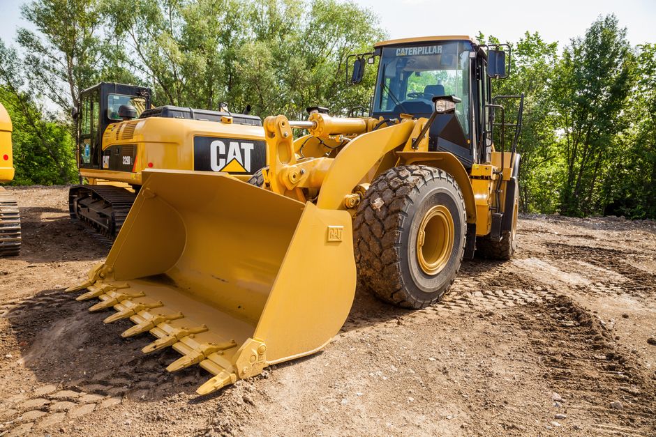 A large yellow Caterpillar front loader construction vehicle parked on a dirt surface with tire tracks, featuring a wide, curved bucket attachment at the front with toothed edges, positioned in an outdoor area surrounded by green trees and a partly cloudy sky. The excavator's cabin with glass windows is visible, along with hydraulic arms connecting the bucket to the main body. In the background, another heavy machinery vehicle is partially visible. The scene appears to be at a construction or excavation site, where heavy equipment is used for earth-moving tasks, and the ground surface shows signs of recent disturbance. Man with Van Wealdstone occasionally uses such equipment during home relocation projects that involve site clearance or access considerations for moving furniture and belongings, aligning with their removal and logistics services.