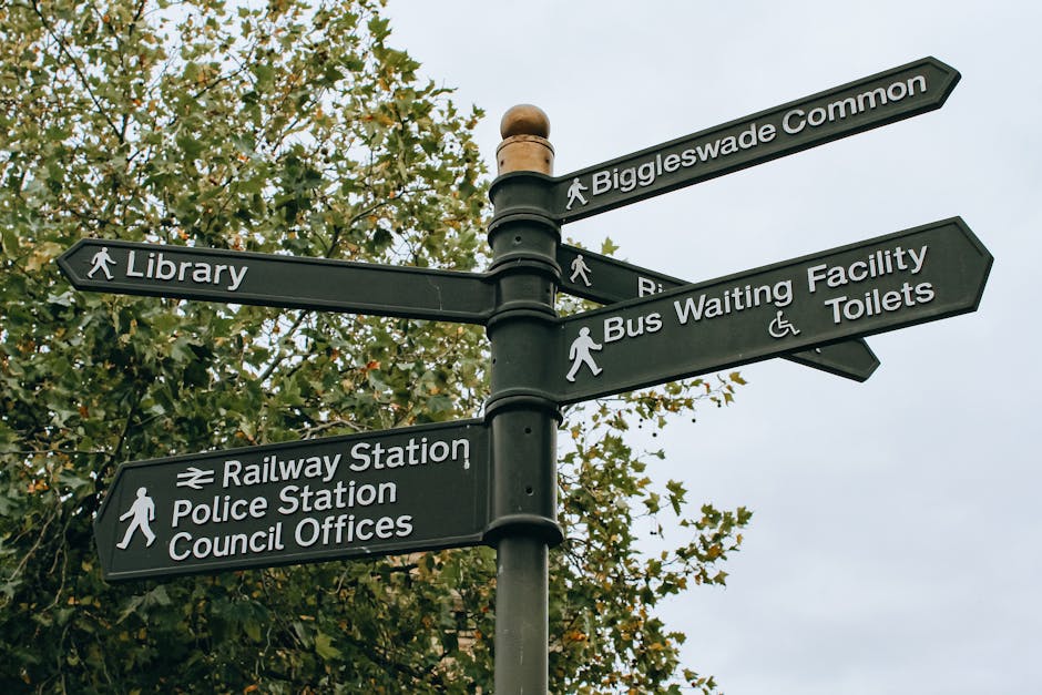 A black multi-directional pedestrian signpost situated outdoors amidst greenery, featuring six arrow-shaped signs pointing in various directions to indicate nearby locations including Biggleswade Common, Library, Railway Station, Police Station, Council Offices, Bus Waiting Facility, and Toilets. The signs are mounted on a central pole with visible fixtures, and each sign displays white text and icons for easy identification. The background includes a tree with green leaves and an overcast sky, suggesting daytime. This type of signage is relevant for guiding residents or visitors during home relocations or local moves in Wealdstone, where clients like Man with Van Wealdstone assist with packing, furniture transport, and moving logistics to comply with local guidelines such as Harrow Council rubbish rules after a move.