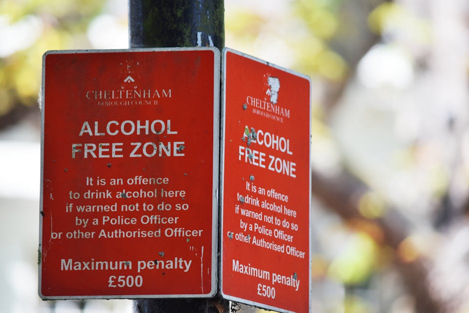 Two red Cheltenham Borough Council alcohol-free zone signs attached to a metal pole outside a residential property. The signs display white text indicating the zone is designated as alcohol free, citing it as an offence to drink alcohol there if not warned to do so by a Police Officer or other authorised personnel, with a maximum penalty of £500. The signs are slightly weathered, with minor scratches and chipped paint, and are positioned on a pavement next to a house entrance, possibly during a home relocation or moving process managed by Man with Van Wealdstone. Blurred autumn foliage in the background indicates an outdoor setting, and the overall scene suggests a street environment where residential or commercial property is being prepared for a move or cleaning, relevant to house removal and transport services.