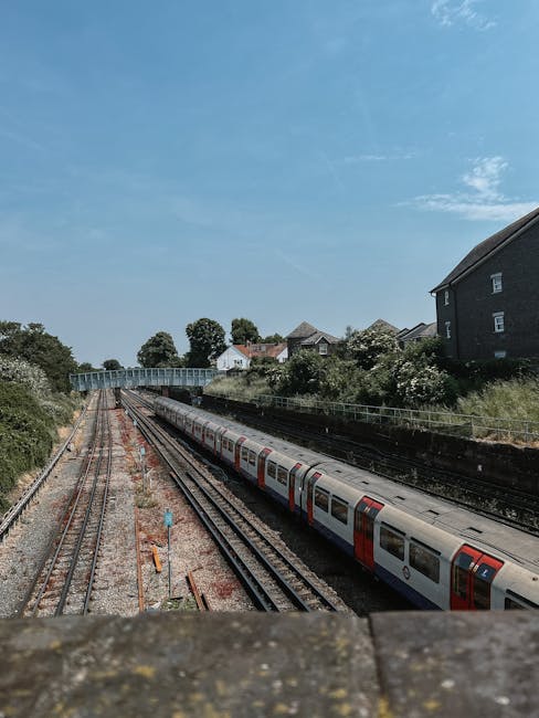 View of a railway station with multiple train tracks extending into the distance under a clear blue sky. A passenger train with a white and red livery is seen on the right set of tracks, partially visible as it moves along the rails. On the left side, adjacent tracks are visible with some orange and blue signalling equipment and wooden sleepers. In the background, a bridge crosses over the tracks, and beyond that, a row of trees and residential houses with dark roofing and brick walls are situated on a slightly elevated area. The scene is typical of an urban or suburban environment suitable for home relocation and moving logistics, with the station’s outdoor setting presented during daytime. Man with Van Wealdstone occasionally provides house removal services, and the image subtly reflects the considerations involved in furniture transport and packing preparations during a moving day process. The overall scene emphasizes the transition point in a move, with focus on the tracks, transportation, and residential surroundings.