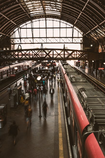 Inside Harrow & Wealdstone station, a large historic railway terminus with a curved, vaulted glass and metal roof allowing natural light to illuminate the platform area. Multiple train tracks run through the station, with a red and grey train stationary on one side. Passengers are walking along the platform, some carrying luggage or backpacks, while others wait near the train doors. In the background, a pedestrian bridge crosses above the tracks, and station signage is visible on the walls. The environment is busy with people moving, indicative of a typical busy transport hub associated with house removals and moving logistics, highlighting the importance of careful planning during furniture transport or packing and moving activities.