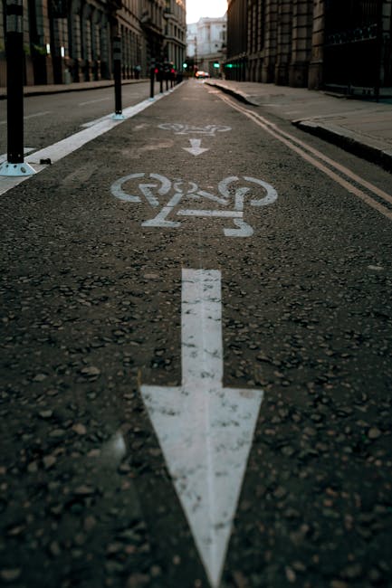 A close-up view of a designated cycling and lane-sharing road marking painted in white on an asphalt street surface. The marking includes a directional arrow pointing straight ahead and a bicycle symbol positioned above it, indicating a cycle lane. The marking is situated in the center of a narrow urban street, with tall, multi-storey buildings lining both sides. Black bollards and posts separate the cycle lane from pedestrian areas on the sidewalk. In the background, traffic lights are visible at an intersection, with faint green signals indicating the direction is clear for movement. The scene is captured in daylight, with soft ambient lighting illuminating the textured asphalt and the road markings, reflecting a typical city street environment suitable for home relocation or transport logistics in a busy urban setting.