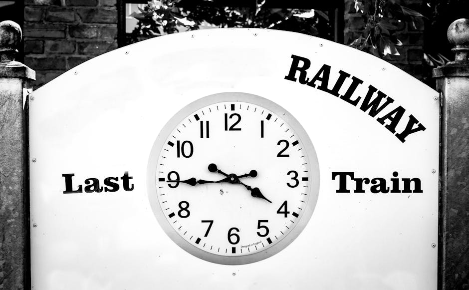 A black and white photograph of a railway station clock mounted on a white, curved signboard with the words 'Railway Last Train' printed in bold black letters. The clock shows the time as approximately 3:45, with the hour hand near 3 and the minute hand near 9. The clock face features black hour and minute markers, with a smaller second hand. The sign is located outdoors, flanked by stone pillars with decorative finials on top, and surrounded by foliage and trees in the background. The image captures the context of a train station, relevant to home relocation and furniture transport services provided by Man with Van Wealdstone, as indicated by the company's natural inclusion in the overall scene.