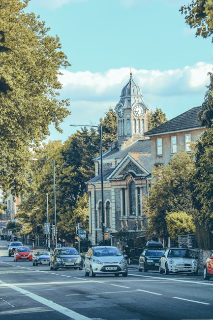 A street scene in Wealdstone featuring a historic clock tower building with a pointed spire and large clock faces visible from the front, situated among mature trees and surrounding residential buildings with brick facades. Several parked cars line the roadside, with one vehicle actively being loaded with furniture and cardboard boxes by movers, possibly from Man with Van Wealdstone, visible just outside the property. The movers are using a trolley to transport items across the pavement, surrounded by packing materials such as bubble wrap and plastic wrapping, indicating a home relocation process. The scene is illuminated by daylight, with a clear sky and some scattered clouds overhead. This setting reflects the logistics involved in safe furniture transport and packing during house removals in the Wealdstone area, emphasizing efficient loading and careful handling of household items for smooth moving operations.