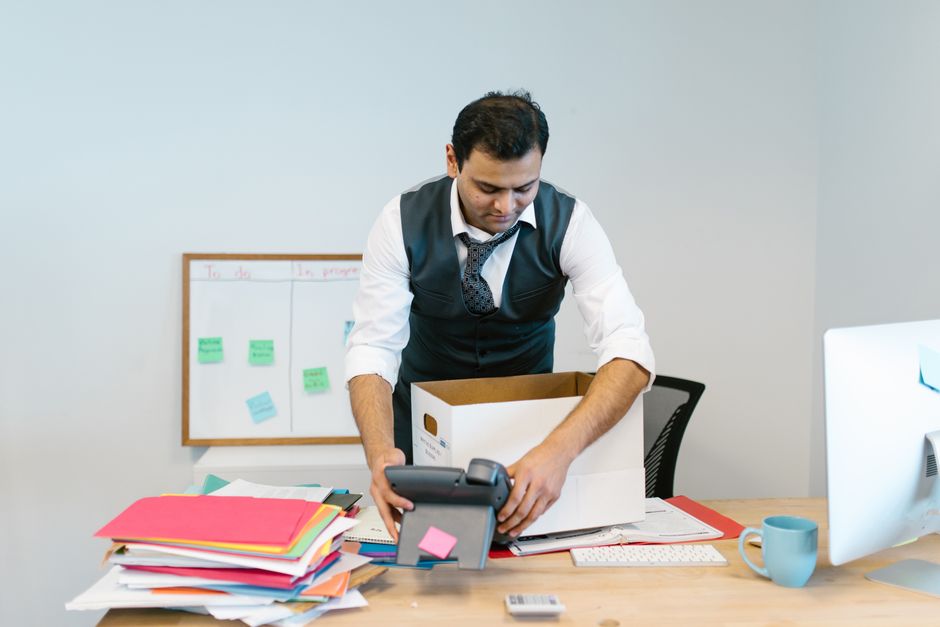 A woman in a beige checkered blazer holding a black storage box containing a potted plant, folders, and documents, standing inside an office environment with modern furniture and glass walls. A man we
