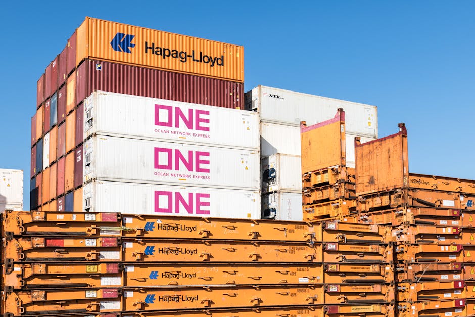A group of stacked shipping containers and flatbed rail car units positioned outdoors against a clear blue sky. The containers include multiple orange Hapag-Lloyd units stacked horizontally at the bottom and middle, with some showing visible branding and identification labels. Above these, there are white containers with pink 'ONE Ocean Network Express' logos stacked vertically, indicating cargo storage for logistics and moving services. The scene depicts a typical setting for freight transportation related to home relocation and furniture transport, illustrating the logistical equipment used in packing and moving processes. The lighting is bright and natural, emphasizing the industrial environment and the scale of equipment managed by companies like Man with Van Wealdstone.