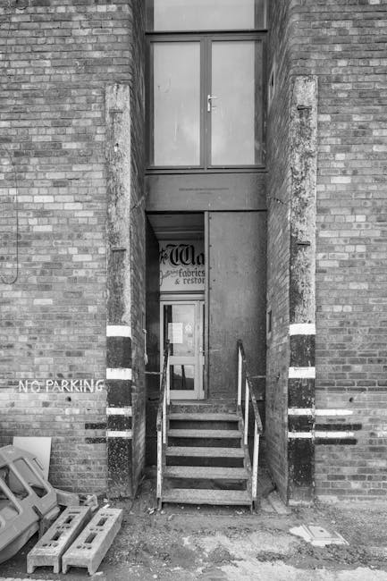 A narrow brick staircase with metal handrails leading up to a second-floor door in a residential building, situated between two brick walls with white painted stripes. The lower part of the staircase is accessible via a small concrete platform, and there are scattered wooden pallets and plastic containers on the ground nearby. In the background, a closed window with a glass pane is visible above the door, and a sign that partially reads 'fabrics & restore' is mounted on the wall behind the stairs. The setting appears to be outside a property, possibly during a home relocation process managed by Man with Van Wealdstone, with a focus on accessing difficult staircases for furniture transport and packing and moving activities.