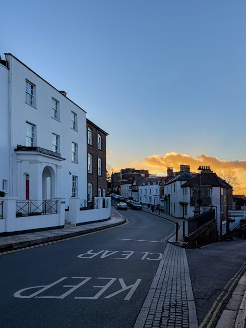 A quiet residential street scene during sunset in Wealdstone with a gentle incline. On the left, there is a large white multi-story building with several windows, a small balcony, and a curved doorway